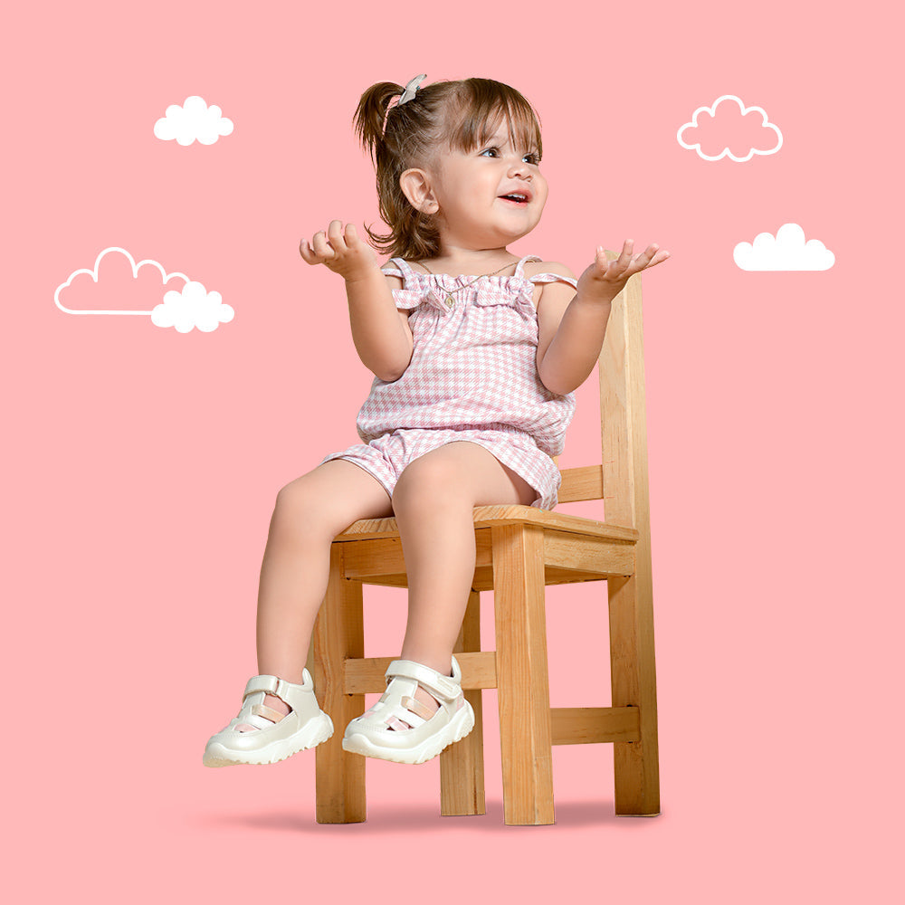 Bebé niña sentada en silla de madera con sandalias blancas coqueta y audaz, y con conjunto rosa a cuadros.