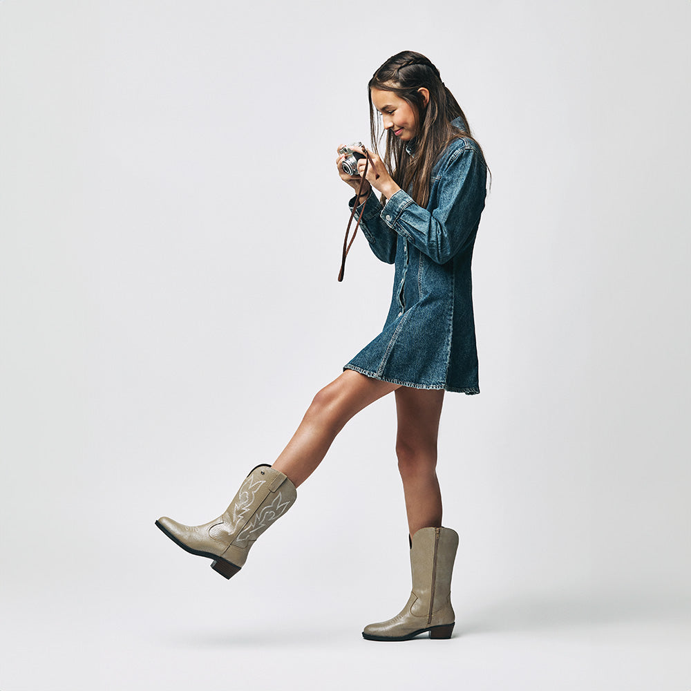 Niña con vestido de mezclilla y botas vaqueras Coqueta y Audaz de piel en color beige, posando con cámara vintage. Moda infantil western que inspira confianza y autenticidad.